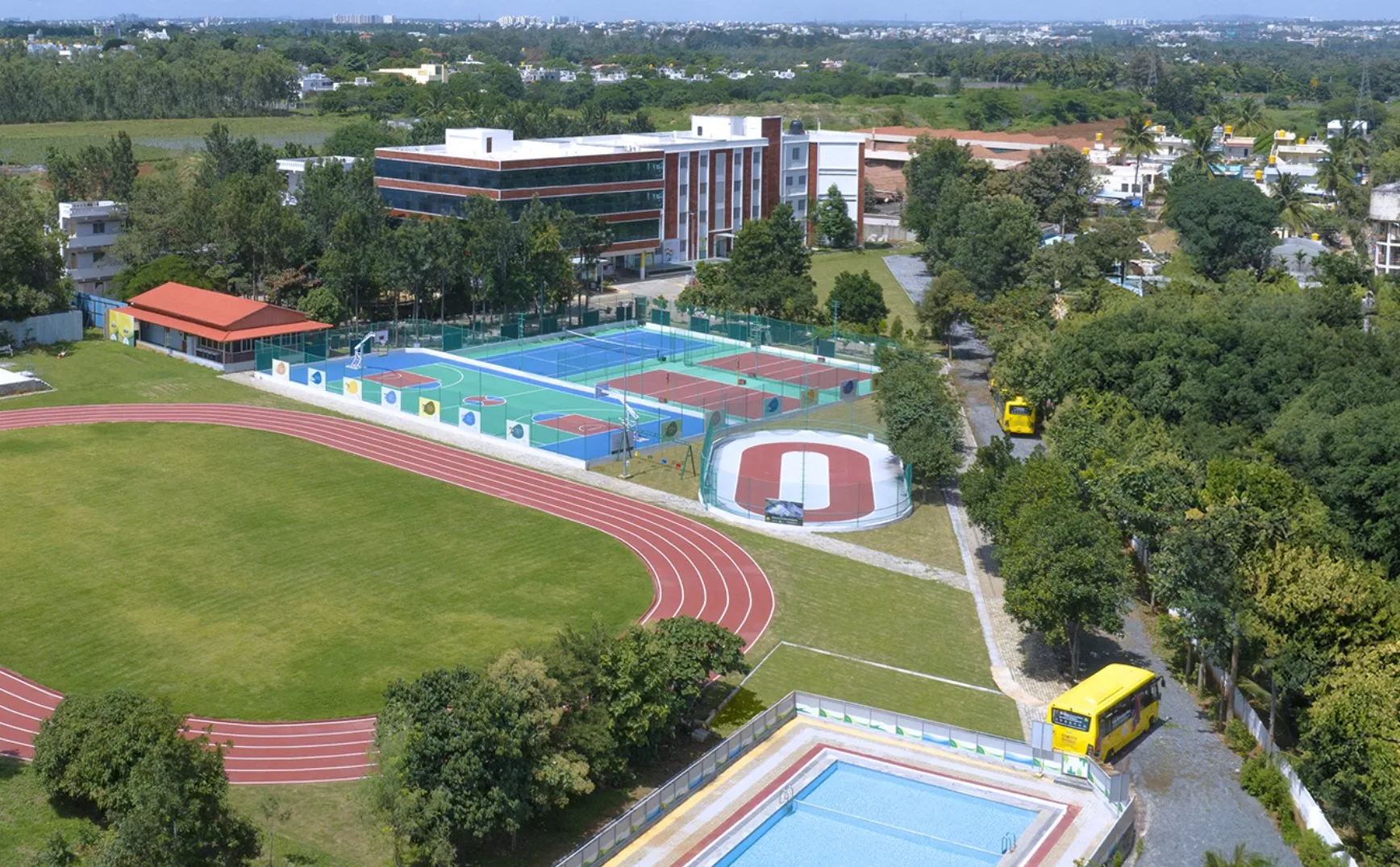 Aerial view of the Electronic City campus: the running track around a grass field, tennis and basketball courts, a skating rink, the swimming pool and the main building behind the trees.